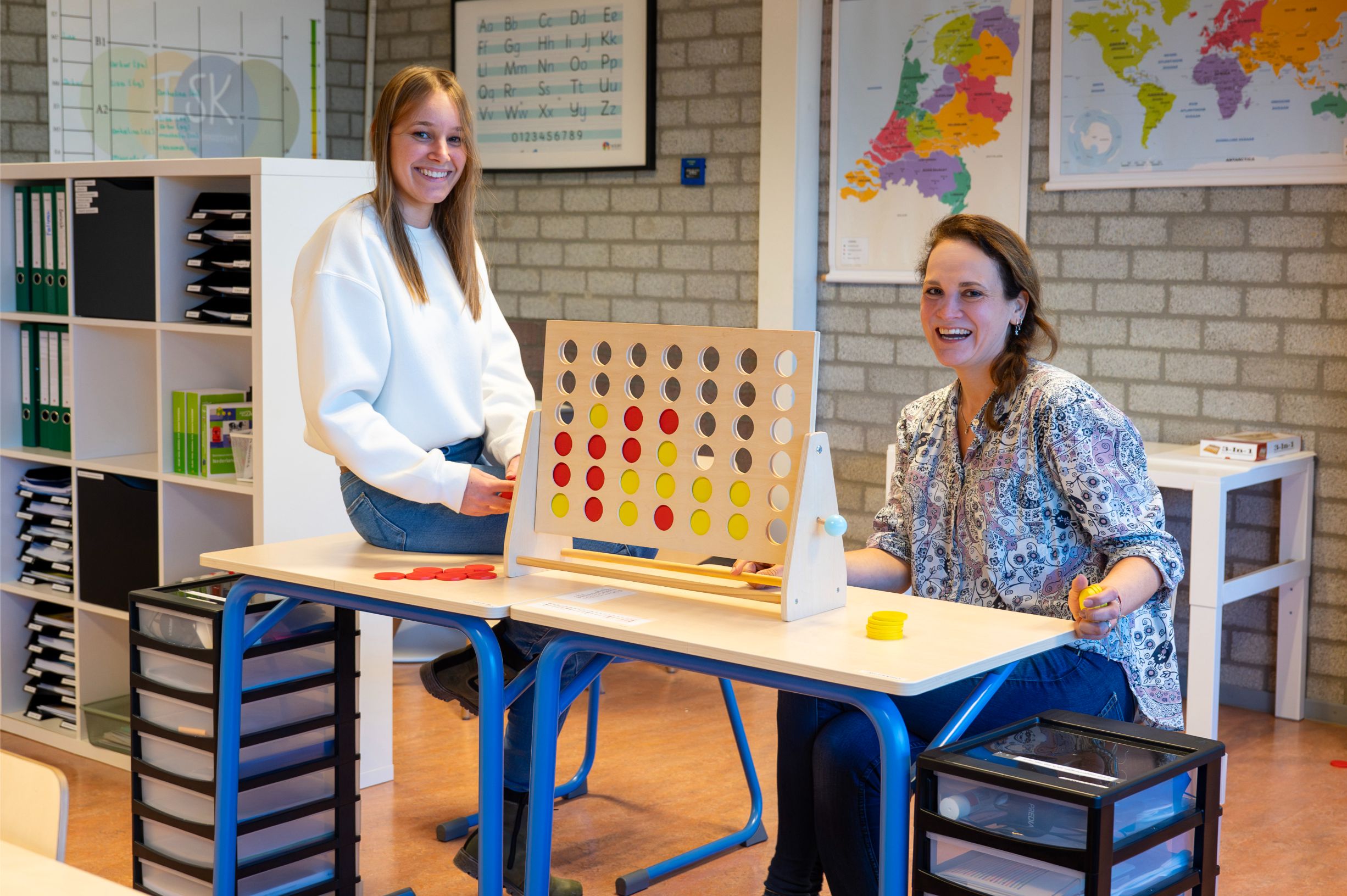Twee vrouwelijke docenten zittend aan schoolbureau met vier-op-een-rij spel voor zich.